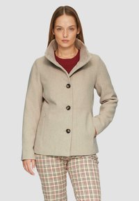 Beżowy płaszcz z wełny z wysokim kołnierzem, z przodem na guziki i bocznymi kieszeniami, sparowany z kraciastymi spodniami w odcieniach czerwieni i beżu.