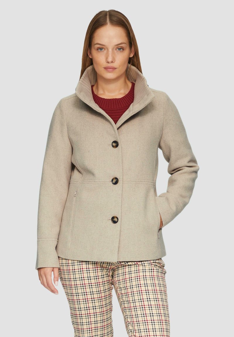 Beżowy płaszcz z wełny z wysokim kołnierzem, z przodem na guziki i bocznymi kieszeniami, sparowany z kraciastymi spodniami w odcieniach czerwieni i beżu.
