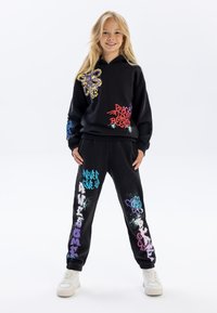 Zwart hoodie en joggingbroek set met kleurrijke graffiti-achtige graphics, met een vlinderontwerp en verschillende teksten in blauw, roze en paars.