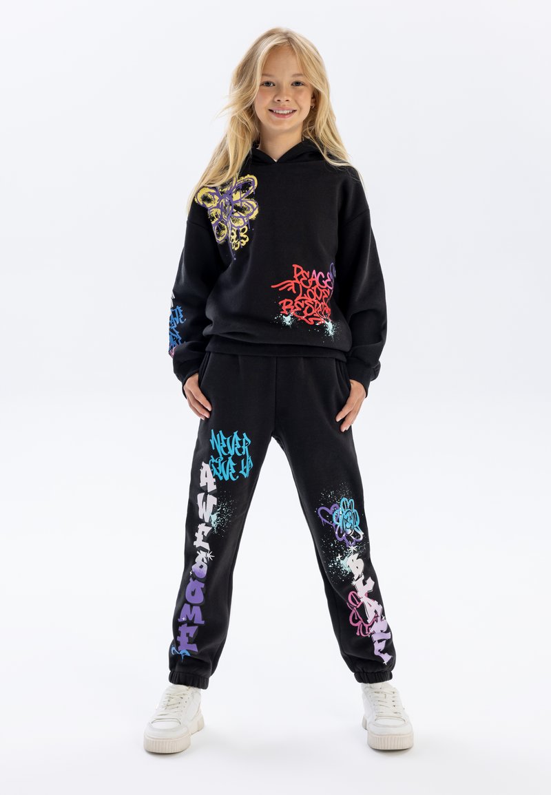 Zwart hoodie en joggingbroek set met kleurrijke graffiti-achtige graphics, met een vlinderontwerp en verschillende teksten in blauw, roze en paars.