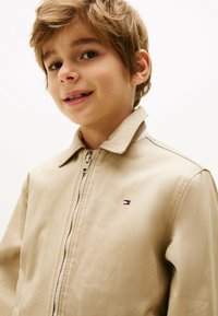 Veste zippée beige avec un col, ornée d'un petit logo sur la poitrine. Texture lisse, design décontracté, adaptée aux temps plus frais.