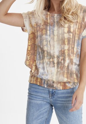 Kobieta ubrana w krótki rękaw, koszulkę tie-dye w ziemistych kolorach i jasnoniebieskie dżinsy, jedna ręka podniesiona, wykadrowana na wysokości ramion i bioder.