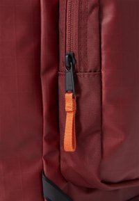 Rode rugzak met een ritsvak met een zwarte rits en een oranje trekhaak. Het materiaal heeft een geprofileerd rasterpatroon.
