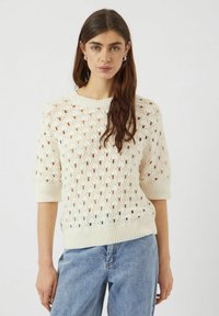 Krämfärgad stickad tröja med en texturerad, öppen spetsdesign, korta ärmar och ribbad krage och nederkant. Parat med ljusblå jeans.