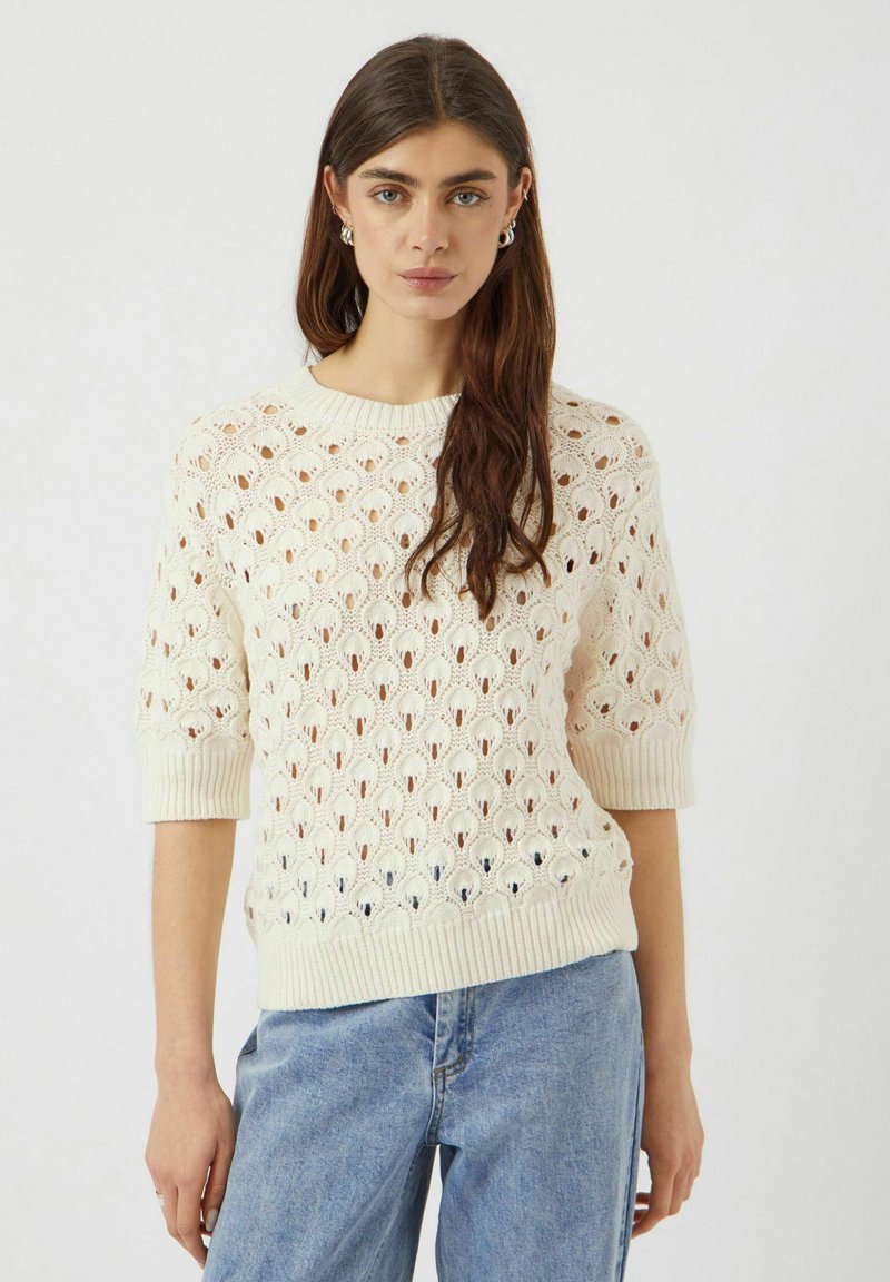 Krämfärgad stickad tröja med en texturerad, öppen spetsdesign, korta ärmar och ribbad krage och nederkant. Parat med ljusblå jeans.