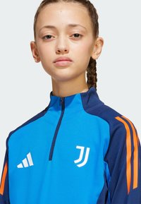 Veste de sport zippée bleu et marine avec des accents orange, présentant un logo et des bandes blanches sur les épaules. Texture lisse, design ajusté.