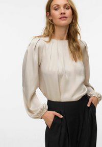 Lysebeige bluse med lange, puffede ermer, rynkede mansjetter og plisserte detaljer, kombinert med høytlivede svarte bukser.