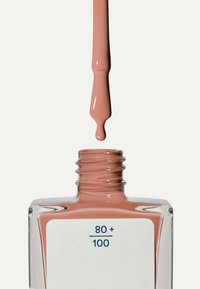Esmalte de uñas marrón claro goteando del pincel en un frasco cuadrado transparente abierto etiquetado "80+ / 100" sobre fondo blanco.
