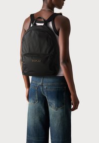 Zaino in nylon nero con logo dorato "REPLAY", dotato di spallacci doppi, manico superiore e tasca frontale con zip, esposto su uno sfondo neutro.