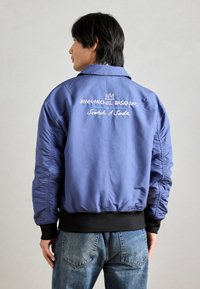 Μπλε μπουφάν bomber φτιαγμένο από λείο ύφασμα, με ριγωτή μαύρη μέση και κεντημένο κείμενο στην πλάτη.