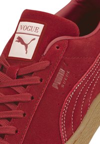 Zapatilla de deporte de ante rojo con superficie texturizada, que presenta un parche blanco con la etiqueta "VOGUE" y el logotipo de Puma, complementada por cordones rojos a juego.