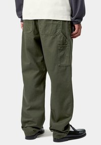 Persona vistiendo pantalones cargo verde oliva de corte holgado, zapatos negros de slip-on y una camiseta de manga larga blanca y gris, de pie de espaldas.