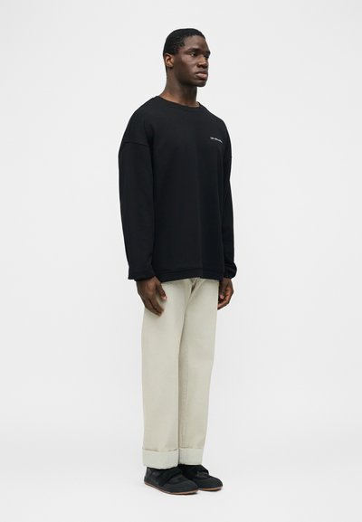 Νεαρός άνδρας στέκεται, φοράει φαρδιά μαύρη μπλούζα με μικρό κείμενο στο στήθος, μπεζ παντελόνι με φαρδιά πόδια και μαύρα παπούτσια, μπροστά σε απλό λευκό φόντο.