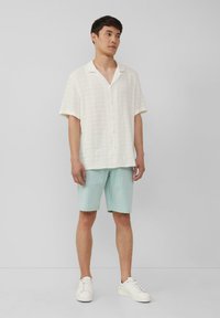 Korte mouw witte textuurshirt met open kraag, gecombineerd met mintgroene shorts en witte sneakers, staand tegen een neutrale achtergrond.
