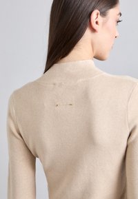 Pulover tricotat bej cu guler înalt, având detalii decorative cu cusături pe spate și accente subtile de aur. Textură netedă.