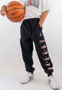 Zwarte trainingsbroek met elastische tailleband, featuring bedrukt "JORDAN" tekst, een logo en zijzakken. In combinatie met witte sneakers en een basketbal.
