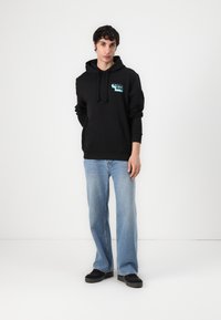 Zwarte hoodie met grafisch opschrift aan de voorkant, trekkoordkap en buidelzak, gecombineerd met lichtblauwe flared jeans en zwarte sneakers.