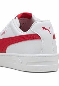 Baskets Puma blanches avec une bande rouge et un logo sur le côté et le talon, montrées de l'arrière et légèrement de profil.