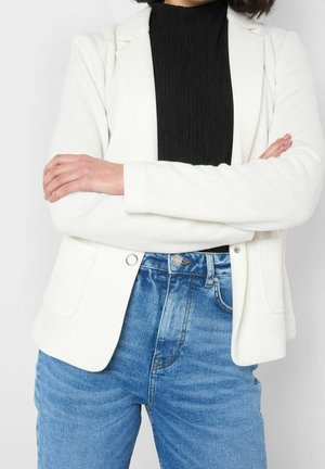 Personne portant un blazer blanc et un top noir côtelé, bras croisés, avec un jean bleu taille haute sur fond uni.