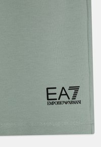 Gaiši zaļš kokvilnas T-krekls ar melnu EA7 Emporio Armani logo kreisajā apakšējā stūrī, ar redzamām šuvju detaļām apakšmalā.
