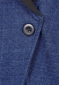 Marinfärgad tyg med en texturerad, prickig mönstring; har en rund svart knapp med fyra små hål som säkrar en flik.