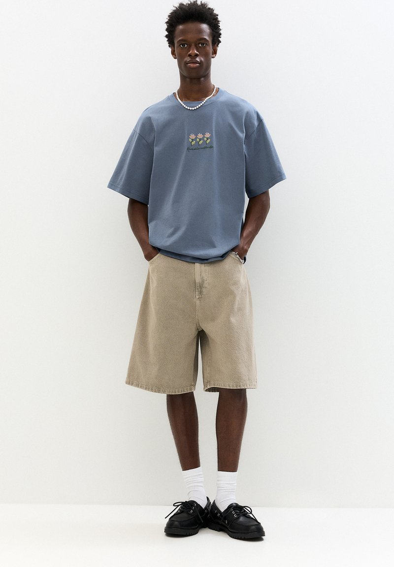 Himmelblaues Baumwoll-T-Shirt mit kurzen Ärmeln und floraler Stickerei, kombiniert mit beigen Denim-Shorts und schwarzen Schuhen, getragen mit weißen Socken.