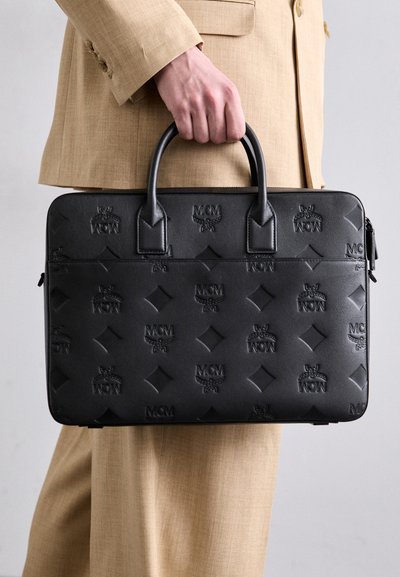 Malette en cuir noir avec logo MCM embossé, de forme rectangulaire, deux poignées et surface texturée. Portée avec un costume beige.