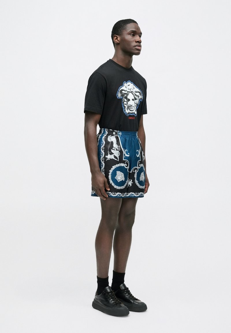 Sort T-shirt med grafisk print og blå mønstrede shorts i letvægtsstof; båret med sorte sneakers og sokker.