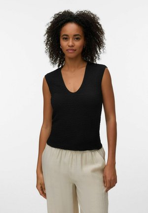 Kvinde med krøllet hår iført en ærmeløs sort tekstureret top og lysebeige løse bukser mod en ensfarvet hvid baggrund.