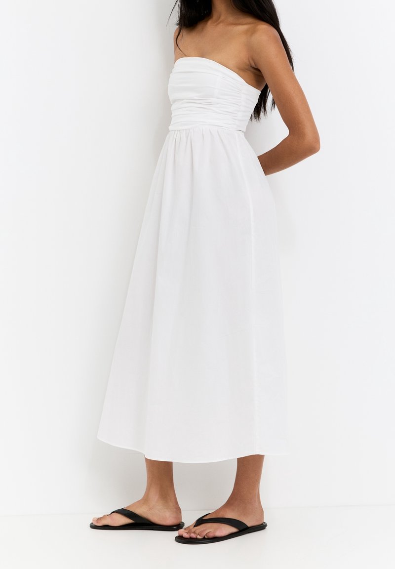 Femme portant une robe midi blanche bustier froncée et sandales plates noires, debout devant un fond blanc.