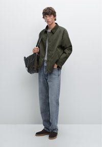 Giacca verde oliva con colletto, camicia grigia sotto, jeans azzurri chiari, scarpe in camoscio marroni e borsa a tracolla nera.