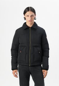 Chaqueta negra acolchada con cierre frontal, dos bolsillos en la parte delantera y un cuello. Presenta detalles cosidos y acentos en bronce. Lleva puesta sobre unos jeans oscuros.