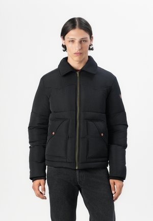 Black padded jacket with a zip front, two front pockets, and a collar. Features stitched detailing and bronze accents. Worn over dark jeans.