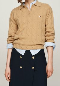 Pull en tricot beige avec col polo, porté par-dessus une chemise à col bleu clair, associé à une jupe marine ornée de boutons dorés.