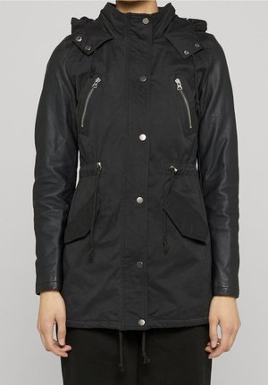 Czarna kurtka parka z srebrnymi zatrzaskami, dwoma zapinanymi na zamek kieszeniami na piersi, klapowymi kieszeniami na wysokości talii, ściągaczami i rękawami o wyglądzie skóry na stojącej osobie.