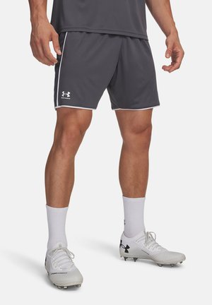 Vīrietis valkā tumši pelēnas Under Armour sporta šortus, baltas sporta zeķes un baltus sporta apavus ar melniem Under Armour logotipiem.