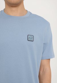 Tricou bleu deschis din bumbac, cu un guler rotund și mâneci scurte. Include un logo brodat rectangular pe partea stângă a pieptului.