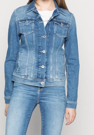 Vrouw draagt een spijkerjasje in medium blauwe wassing met knoopsluitingen en bijpassende spijkerbroek, tegen een neutrale achtergrond.