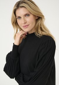 Schwarzer Pullover mit hohem Kragen und plissierten Ärmeln. Glatte Textur mit tailliertem Design, das sanfte, fallende Akzente betont.