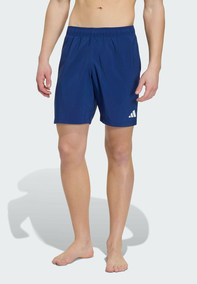 Modèle masculin portant un short de sport bleu avec le logo Adidas sur la jambe gauche, debout pieds nus sur un fond clair uni.