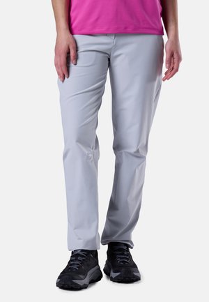 Person iført lysegrå bukser med lige ben, sorte vandresko og en lyserød top, stående med hænderne afslappet ved siden.