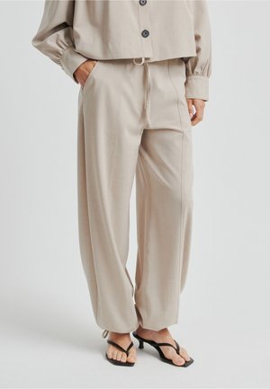Beżowe spodnie na miarę o luźnym kroju, z zakładkami i regulowanym sznurkiem. Lekka tkanina, boczne kieszenie, w zestawieniu z czarnymi klapkami.