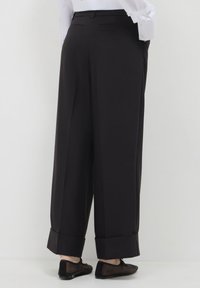 Persona che indossa pantaloni neri a gamba larga con risvolti, camicia bianca infilata dentro, e scarpe flat nere, in piedi davanti a uno sfondo bianco uniforme.