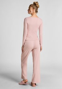 Vrouw met blond haar in een knot, gekleed in een bijpassende roze lange mouwen top en broek met roze pluizige slippers, staand met de rug naar ons toe.