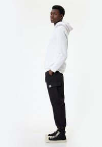 Hvid hættetrøje, sorte cargo-bukser med sidelommer og sorte højhalsede sneakers. Glat tekstur, afslappet pasform, minimalistisk design.