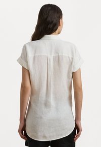 Short-sleeve white linen shirt with a back yoke, soft texture, and rolled cuffs. Features a curved hem and subtle translucence.