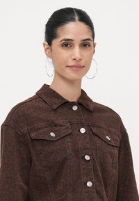 Bruine denimjas met een luipaardprintpatroon, knoopsluiting aan de voorkant, twee borstzakken en een puntige kraag. Zilveren drukknoopjes accentueren het ontwerp.
