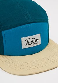 Kappe mit einem teal- und blauen Stoffkörper, einem beigefarbenen Schirm und einem rechteckigen weißen Etikett, auf dem "Lil' Boo Copenhagen" in schwarzer Schrift steht.
