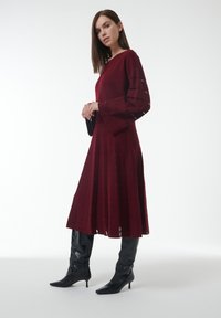 Robe en tricot bourgogne à manches longues, présentant des découpes verticales et une jupe plissée, assortie à des bottes noires jusqu'aux genoux.