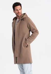 Beige hættefrakke lavet af glat stof, med lynlåslukning, snørredetaljer og sidelommer, parret med lyseblå bukser.